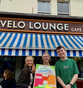 Staff standing outside Velo Lounge holding a colouful board with some of the activities they have planned for Lounge Aid